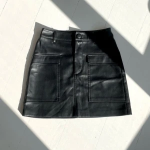 Svart skinnkjol från Zara - Snygg svart skinnkjol från Zara med stora fickor framtill och en knappdetalj i midjan. Kjolen har en stilren design och är perfekt till middag som utgång 💗