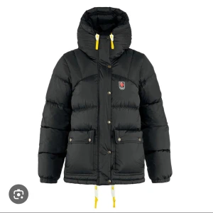 Svart dunjacka från Fjällräven - Säljer en svart dunjacka från Fjällräven med gula detaljer. Jackan har en hög krage och dragkedja med knappar framtill. Två stora fickor med knappar och justerbara snören i nederkant. Perfekt för kalla dagar!