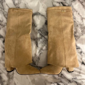 Beige mockastövlar  - Snygga beige stövlar i mocka med spetsig tå och klack. De har en dekorativ rem med guldfärgat spänne upptill och en praktisk dragkedja på insidan för enkel på- och avtagning. Helt nya och oanvända. Skulle gissa på att klackhöjden är 7cm. Nypris: 699kr