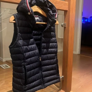 Svart dunväst från Tommy Hilfiger - Säljer en snygg svart dunväst från Tommy Hilfiger i storlek XS. Västen har en praktisk dragkedja och en stilren design med huva. Perfekt inför vår och sommaren 