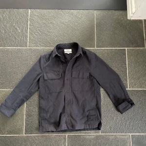 Marinblå overshirt från COS - Snygg marinblå overshirt från COS med långa ärmar och knappar framtill. Perfekt för lager-på-lager stil. Två praktiska bröstfickor ger en cool touch. Passar både till vardags och mer uppklädda tillfällen.