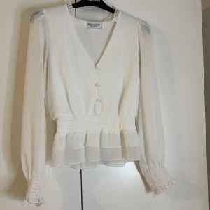 Vit fräsch blus med snygga detaljer  - Jag säljer en vit snygg blus med snygga detaljer och resår i midjan och ärmarna en av knapparna är lite lösa och i bild tre visar jag ena armen där resåren gått upp lite men annars nästan i helt nytt skick😊