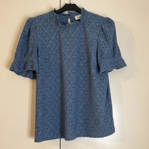 Blå blus med broderad design - Säljer en söt blå blus från Vila med broderad design och korta ärmar. Blusen har en rund hals och en knappdetalj i nacken. Perfekt för en somrig look!