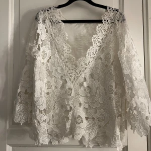 Vit spetsblus med blommönster - Elegant vit spetsblus med vackert blommönster. Blusen har trekvartsärmar och en djup v-ringning framtill. Perfekt för en stilfull look.