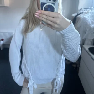 Vit stickad tröja med knytband - Säljer en stilren vit stickad tröja med långa ärmar och dekorativa knytband på sidorna. Perfekt för en mysig och avslappnad look. Tröjan har en normal passform och är idealisk för kyligare dagar.