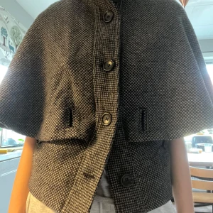 Grå kappa med knappar - Snygg grå kappa med ett diskret rutigt mönster och stora knappar framtill. Jackan har en unik design med kortare ärmar och en elegant passform. Perfekt för att ge din outfit en stilfull touch!! 