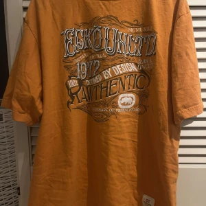 Orange t-shirt från Ecko Unltd - Säljer en cool orange t-shirt från Ecko Unltd med ett stort tryck på framsidan. T-shirten är kortärmad och gjord av bomull, perfekt för en avslappnad stil. Storleken är XL och den har en klassisk passform. Den är köpt i USA.