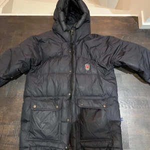Svart dunjacka från Fjällräven - Säljer en svart dunjacka från Fjällräven med huva och praktiska fickor framtill. Jackan har en dragkedja och knappar för extra skydd mot kyla. Perfekt för kalla vinterdagar. 