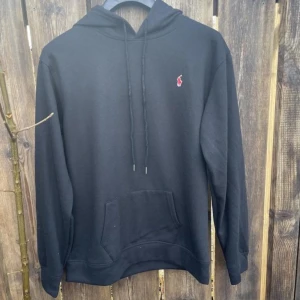 Svart hoodie från Ralph Lauren - Säljer en stilren svart hoodie från Ralph Lauren med det klassiska röda logotypen på bröstet. Tröjan har en praktisk magficka och justerbar huva med snören. Perfekt för en avslappnad look.