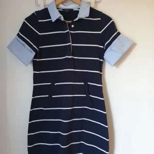 Klänning - Snygg randig klänning i marinblått och vitt med ljusblå krage och ärmslut. Klänningen har korta ärmar och knappar på baksidan. Perfekt för en stilren look! Märket är Gant och den är i mycket bra skick