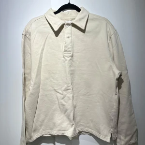 Beige långärmad pikétröja - East West - Säljer en stilren beige långärmad pikétröja med klassisk krage och knappar framtill. Perfekt för en avslappnad men ändå snygg look. Passar bra till både jeans och chinos. Från East West. Lite croppad look.