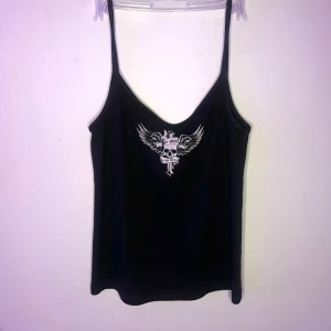 Svart linne  - Snyggt svart linne från Brandy Melville med smala axelband och ett coolt dödskalletryck med vingar på framsidan. Perfekt för en edgy look.