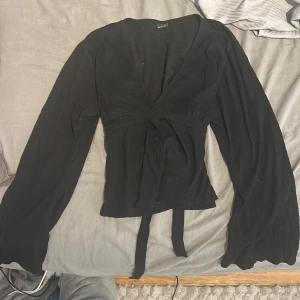 Svart omlottopp från Gina Tricot - Säljer en elegant svart omlottopp från Gina Tricot. Toppen har långa ärmar och ett knytband i midjan för en smickrande passform. Perfekt för en stilren look.