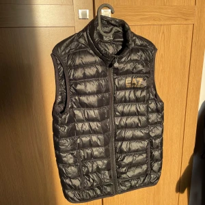 Svart dunväst från EA7 - Snygg svart dunväst från EA7 med dragkedja och hög krage. Perfekt för kyliga dagar och ger en sportig look. Västen har ett diskret EA7-märke på bröstet. Lite små hål på ryggen och ett litet på magen.