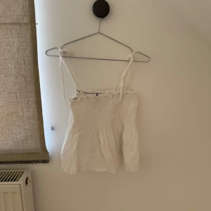 Vit ärmlös topp - Säljer en söt vit ärmlös topp med knytband på axlarna. Toppen har en smockad överdel och en lätt volang nedtill, vilket ger en fin passform. Perfekt för varma sommardagar!
