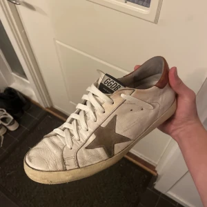 Golden goose skor - Säljer ett par vita sneakers från Golden Goose med en distinkt stjärndetalj i grått på sidan. Skorna har bruna detaljer vid hälen och klassisk snörning. Perfekta för en avslappnad stil. De är slitna men går väl att använda. Självklart äkta och allt på sista bilden tillkommer.