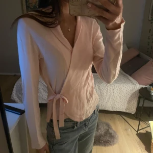 Rosa omlottopp - Säljer en söt rosa omlottopp med knytband i midjan. Toppen har långa ärmar och en v-ringad design som ger en elegant touch. Perfekt för en avslappnad men stilren look.