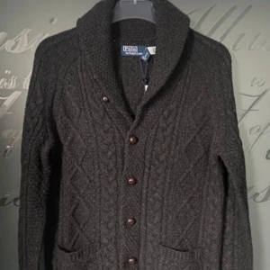 Mörkgrå stickad kofta från Ralph Lauren - Säljer en snygg mörkgrå stickad kofta från Ralph Lauren med flätmönster och knappar framtill. Koftan är sprillans ny och lapparna är kvar. Koftan har en hög krage och praktiska fickor. Perfekt för kyliga dagar!  Ny pris 3600kr