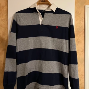 Randig pikétröja från Ralph Lauren - Säljer en randig pikétröja från Ralph Lauren i marinblått och grått. Storlek M. Tröjan har ett litet hål uppe vid ryggen/axeln men detta syns inte när man har på sig den. 