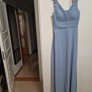 Ljusblå långklänning med spetsdetaljer - Elegant ljusblå långklänning med v-ringad hals och vackra spetsdetaljer på axelbanden. Klänningen har en draperad överdel och ett flytande fall som ger en romantisk känsla. Perfekt för speciella tillfällen.