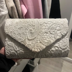 Vit spetskuvertväska - Elegant vit kuvertväska täckt med vacker spets.. Väskan har en praktisk magnetknäppning och ett rymligt inre fack för dina nödvändigheter⚡️ fläckar förekommer på baksida (se sista bild) men det är inget som är särskilt synligt (syns endast i vissa ljus)