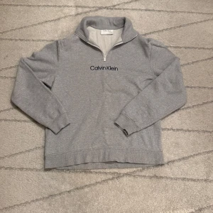 Grå tröja med dragkedja från Calvin Klein (Mycket bra skick) - Säljer en stilren grå tröja från Calvin Klein med halv dragkedja och broderad logga på bröstet. Perfekt för en avslappnad look.