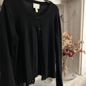 Svart kofta från H&M - Säljer en stilren svart kofta från H&M. Den har långa ärmar och knappar framtill. Perfekt för lager-på-lager-stil eller som ett enkelt basplagg i garderoben. Använd 1 gång så inga tecken på använd! Inga fläckar eller hål!💌💌i storlek s, 200kr! Kom privat för bild med den på.