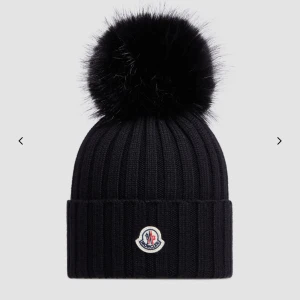 Svart ribbad mössa med pälsboll från Moncler - Snygg svart ribbad mössa från Moncler med en stor pälsboll på toppen. Mössan har en uppvikt kant och Monclers logotyp framtill. Perfekt för kalla dagar!