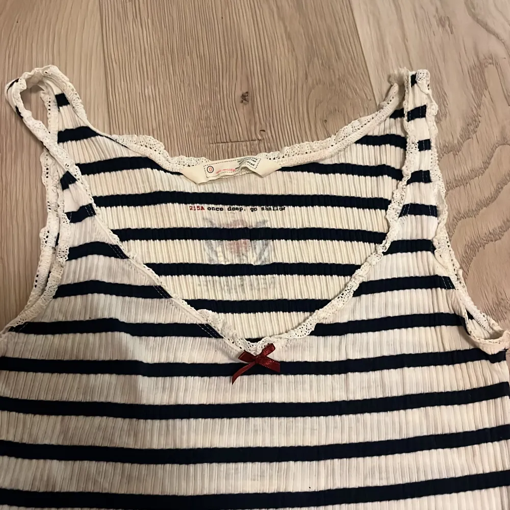 Säljer ett randigt linne i vitt och blått med spetsdetaljer vid axelbanden och en liten rosett framtill. Linnet har en broderad text 'Rise Above' på baksidan. Supergulligt. Passar XS-S. Yläosat.