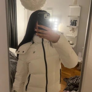 Calvin Klein jacka - Säljer en snygg vit dunjacka med hög krage och dragkedja. Jackan har en stilren design med svarta detaljer och praktiska fickor. Perfekt för kyliga dagar.