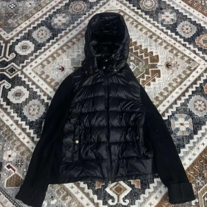 Svart pufferjacka från Moncler - Snygg svart pufferjacka från Moncler med quiltad design och huva. Jackan har långa ärmar i stickat material och en dragkedja framtill. Perfekt för kyligare dagar.