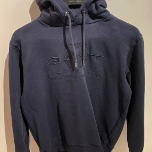 Mörkblå hoodie från GANT - Säljer en snygg mörkblå hoodie från GANT. Tröjan har en klassisk design med justerbar huva och ribbade muddar vid ärmslut och nederkant. Perfekt för en avslappnad stil. 