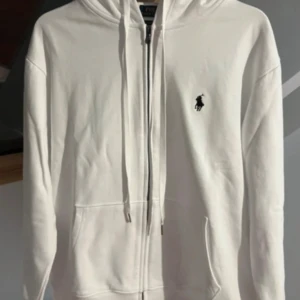 Vit hoodie från Ralph Lauren - Säljer en stilren vit hoodie från Ralph Lauren med dragkedja och det klassiska logotypbroderiet på bröstet. Hoodien har en justerbar huva med snören och praktiska fickor framtill. Perfekt för en avslappnad och snygg look.