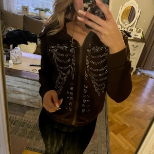 Svart hoodie med skelettmönster - Cool svart hoodie med glittrigt skelettmönster på framsidan. Den har en dragkedja och långa ärmar, perfekt för en unik stil.