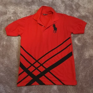 Röd pikétröja från Ralph Lauren - Snygg röd pikétröja från Ralph Lauren med svart logotyp på bröstet och ett coolt rutmönster i svart. Perfekt för en stilren look med korta ärmar och klassisk krage.