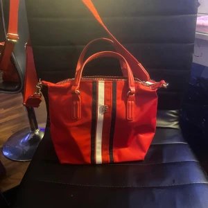 Röd axelväska med randigt mönster - Snygg röd axelväska med vita och svarta ränder. Väskan har en praktisk dragkedja och justerbar axelrem. Perfekt för att ge en färgklick till din outfit.