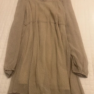 Beige prickig klänning - Charmig beige klänning med små prickar och långa ärmar. Perfekt för en stilren och avslappnad look. Klänningen har en luftig passform och är tillverkad i ett lätt material.