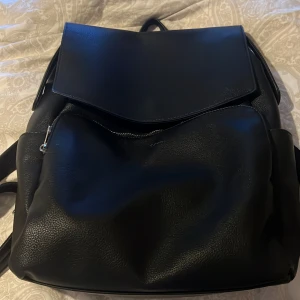 Svart ryggsäck från Rizzo - Snygg och praktisk svart ryggsäck från Rizzo i skinn. Väskan har en stilren design med ett stort lock och dragkedja för säker förvaring. Insidan är rymlig med flera fack för enkel organisering. Perfekt för både vardag och resa.väskan är helt ny och har inga defekter