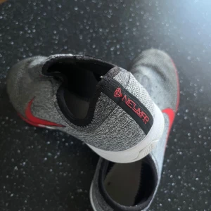 Neymar Futsal skor storlek 40  - Snygga grå och röda Nike sneakers med en stilren design. Skorna har en röd Nike-logga på sidan och en röd sula som ger en sportig look. Perfekta för både träning och vardag. Det finns  defekter men inte något man ser tydligt.. Se på bild 4 och 5. Skorna är annars i mycket bra skick.