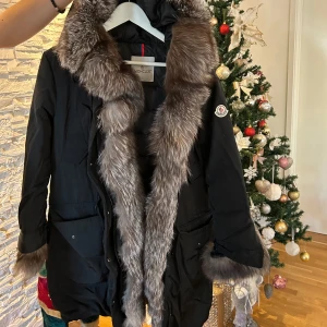 Svart dunjacka med pälsdetaljer från Moncler - Snygg svart dunjacka från Moncler med lyxiga pälsdetaljer på huva och ärmslut. Jackan har en klassisk design med lång ärm och är perfekt för kalla vinterdagar. Den är tillverkad i högkvalitativt material och har en elegant look. Väldigt lite använd, funkar som en S/M