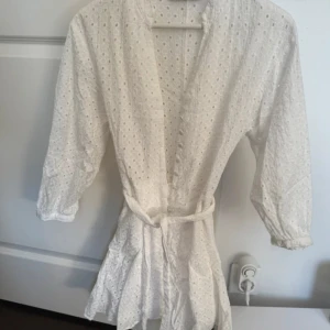 Vit klänning från Zara - Charmig vit klänning från Zara med broderade detaljer och knappar framtill. Klänningen har trekvartsärmar och ett knytband i midjan för en smickrande passform. Perfekt för en somrig look!