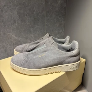 Grå sneakers från Axel Arigato - Snygga grå sneakers från Axel Arigato i mocka med en stilren design. Skorna har en diskret logga på sidan och en bekväm sula i vitt. Perfekta för en avslappnad stil.