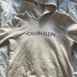Beige hoodie från Calvin Klein - Säljer en stilren beige hoodie från Calvin Klein med svart logotyp på bröstet. Perfekt för en avslappnad look. Hoodien har en klassisk passform och är tillverkad i mjukt material.