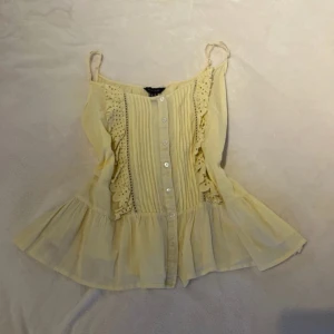 Gul blus från New Look - Charmig gul blus från New Look med spetsdetaljer och knappar framtill. Den har smala axelband och en luftig passform som ger en romantisk känsla. Perfekt för varma dagar. Storlek L men passar mig fint som bär S.