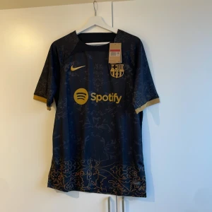 Svart och guld fotbollströja från Nike - Snygg svart fotbollströja från Nike med guldiga detaljer och klubbmärke. Tröjan har korta ärmar och ett elegant mönster. Perfekt för fotbollsfans som vill ha något unikt och stilrent. Barcelonaborna tröja