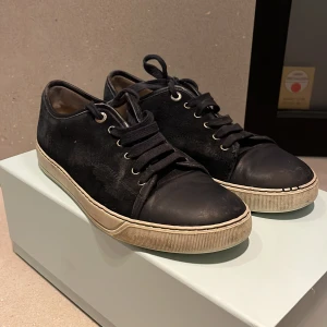 Svarta sneakers från Lanvin - Snygga svarta sneakers från Lanvin med klassisk design. Skorna har en ovandel i skinn och snörning framtill. Tillverkade i Portugal, dessa sneakers kombinerar stil och komfort perfekt. Skorna har lite skador i sulan högra skon med också lite skin som e sönder som man ser på 5 bilden. Pris kan diskuteras. Kommer med orginal box