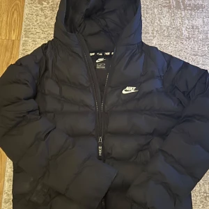 Svart dunjacka från Nike - Säljer en svart dunjacka från Nike med huva och dragkedja. Jackan har en klassisk pufferdesign och är perfekt för kyligare väder. Den har en diskret Nike-logga på bröstet.