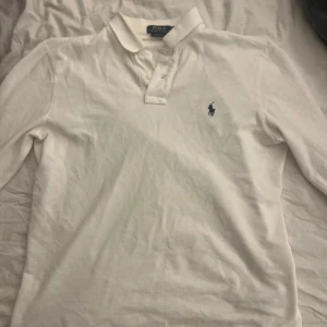 Vit pikétröja från Polo Ralph Lauren - Säljer en klassisk vit pikétröja från Polo Ralph Lauren med den ikoniska loggan broderad i blått på bröstet. Tröjan har långa ärmar och en stilren krage med knappar. Perfekt för en avslappnad men ändå stilren look.