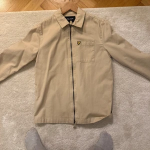 Beige overshirt från Lyle & Scott - Snygg beige overshirt från Lyle & Scott med dragkedja och en broderad logga på bröstet. Jackan har en klassisk krage och långa ärmar, perfekt för vår och höst. Storlek S. Den är knappt använd och är i mycket bra skick