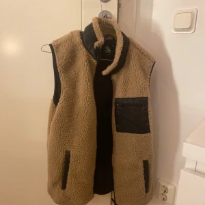 Beige fleeceväst med svarta detaljer - Säljer en mysig beige fleeceväst med svarta detaljer. Västen har en hög krage och praktiska fickor fram. Perfekt för kyliga dagar!